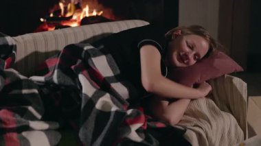 Genç kız şöminenin yanındaki kanepede huzur içinde uyuyor. Sıcak bir Noel akşamında ekose bir battaniyeye sarılı. Yüksek kalite 4k görüntü