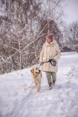 Karlı kırsal bir yolda Golden Retriever 'ıyla yürüyen yaşlı bir kadın. Kadın, sakin bir kış gününde etrafı karla kaplı ağaçlarla çevrili tasmayı tutarken köpeğe sıcak bir şekilde gülümsüyor. Yüksek