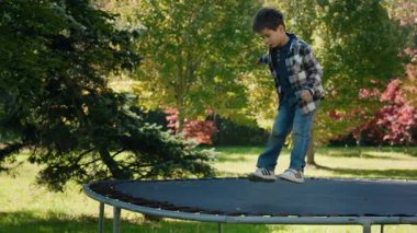 Yeşil ağaçlar ve sonbahar renkleriyle çevrili bir trambolinde zıplayan mutlu çocuk dışarıda aktif bir günün tadını çıkarıyor. Yüksek kalite 4k görüntü