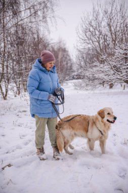 Mavi kışlık ceketli yaşlı bir kadın, elinde Golden Retriever 'ıyla karlı bir yolda duruyor. Kadın düşünceli bir şekilde karla kaplı ağaçların arasına bakarken tasmalı köpek kış havasının tadını çıkarıyor. Yüksek