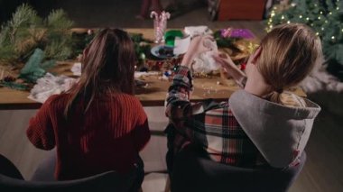 İki kişi bir masada oturmuş çam dalları, takılar ve şenlikli ışıklarla çevrili Noel süslemeleri yaparak sıcak bir tatil ortamı yaratıyor. Yüksek kalite 4k görüntü