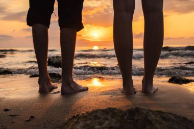 Gün doğumunda kumlu sahilde çıplak ayaklı çift denizdeki dalgaları ve altın yansımaları seyrediyor. Yüksek kalite fotoğraf