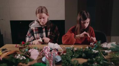 İki genç kadın, şöminenin yanındaki neşeli çelenkler, süsler ve şeker kamışlarıyla dolu rahat bir masada Noel süsü hazırlıyorlar. Yüksek kalite 4k görüntü