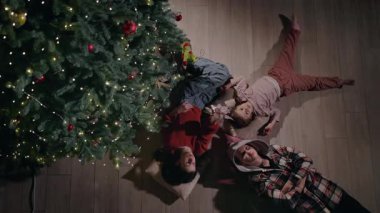 Aile, dekore edilmiş bir Noel ağacının altında yerde dinleniyor sıcak ışıklar ve sıcak bir atmosferle evde huzurlu bir bayram akşamının tadını çıkarıyor. Yüksek kalite 4k görüntü
