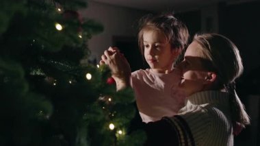 Genç bir kızı Noel ağacına kırmızı bir süs asarken tutan bir kadın. Ev ortamında parıldayan ışıklarla süslenmiş. Yüksek kalite 4k görüntü