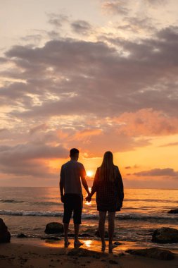 Çiftler el ele tutuşup deniz kenarında güneşin doğuşunu seyrederler. Sahilde çıplak ayakla dururlar. Sıcak güneş ışığı suya vurur. Yüksek kalite fotoğraf