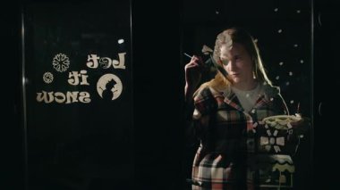 Genç bir kadın gece pencereye kardan adam ve neşeli kar taneleri çiziyor. 