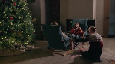 Anne ve iki genç kızı Noel ağacının yanında oturmuş, sıcak ve şenlikli ışıklarla dolu sıcak bir ev atmosferinde birlikte örgü örüyorlar. Yüksek kalite 4k görüntü