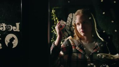 Geceleri pencereye şenlikli tasarımlar çizen genç bir kadın, evin içinde sıcak bir ışık ve arka planda parlayan Noel süslemeleriyle aydınlatılıyor. Yüksek kalite 4k görüntü