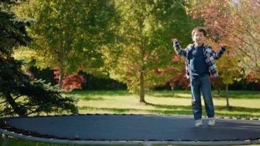 Güneşli bir günde trambolinde zıplayan gülümseyen çocuk. Etrafı renkli ağaçlar ve yeşil çimlerle çevrili. Yüksek kalite 4k görüntü