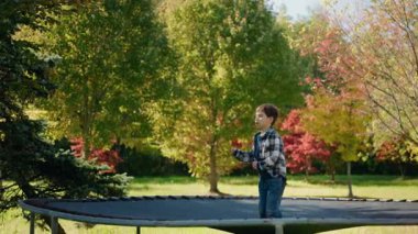 Küçük çocuk yeşil ve renkli sonbahar ağaçlarıyla çevrili güneşli parkta trambolinde zıplıyor. Yüksek kalite 4k görüntü