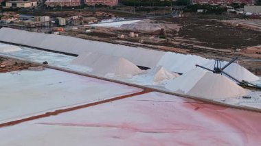 Gün batımında pembe tuzlu su lagünlerinin yakınındaki büyük beyaz tuz yığınlarının hava görüntüsü, taşımacılık makineleriyle endüstriyel tuz üretimini gösteriyor. Yüksek kalite 4k görüntü