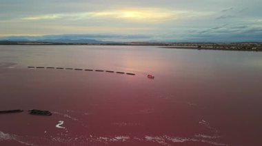 Gün batımında Torrevieja Tuz Gölü 'nün pembe sularında çekilen küçük bir sanayi teknesinin insansız hava aracı görüntüsü. Dağlar ve ufukta görünen şehir. Yüksek kalite 4k görüntü
