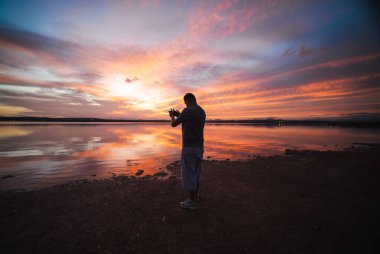 Gün batımında bir dron kontrolörüyle kıyıda duran adam, sakin göl yüzeyinde turuncu ve mor bulutların güzel yansımaları. Yüksek kalite fotoğraf