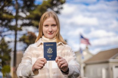 Dışarıda duran genç bir kadın, ellerini ileri uzatıyor ve elinde banliyö evleri ve arka planda Amerikan bayrağı olan mavi bir ABD pasaportu var. Yüksek kalite fotoğraf