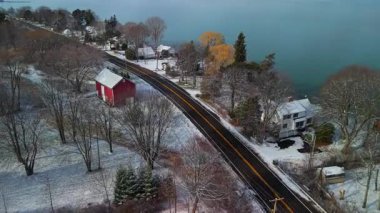 Karlı ağaçların, kırmızı bir ahırın ve kıyı şeridi boyunca yerleşim yerlerinin yanından geçen göl kenarındaki bir yolun hava manzarası. Yüksek kalite 4k görüntü