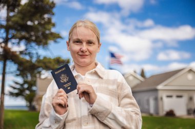 Arka planında Amerikan bayrağı olan, Amerika Birleşik Devletleri pasaportu taşıyan gülümseyen bir kadın vatandaşlığı, göçmenlik ve seyahat özgürlüğünü simgeliyor. Yüksek kalite fotoğraf