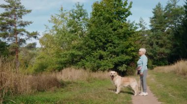 Yeşil parktaki toprak yolda Golden Retriever köpeğiyle ayakta duran yaşlı kadın. Yüksek kalite 4k görüntü