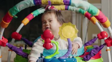 Bebek kız renkli bir kazakla neşeli bir şekilde zıplıyor, etrafındaki parlak oyuncakları keşfederken komik bir ifade takınıyor. Yüksek kalite 4k görüntü