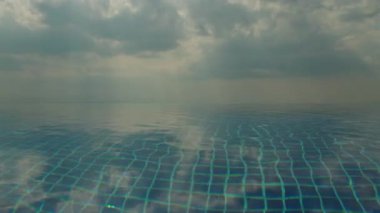 Sakin sonsuz havuz su yüzeyi yumuşak yansımaları ve bulutlu gökyüzü, mavi fayanslı asgari soyut sahne, sakin ruh hali ve büyük kopya alanı. Yüksek kalite 4k görüntü