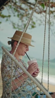 Orta yaşlı bir kadın tropik sahilde iple sallanan sandalyede oturmuş, huzur dolu deniz manzarasının tadını çıkarırken smoothie içiyor. Yüksek kalite 4k görüntü