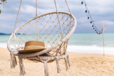 Hasır şapka, yumuşak okyanus dalgaları ve sicim ışıklarıyla kumlu sahilde sallanan halat sandalyesinde dinleniyor, sakin deniz kenarı rahatlama sahnesi. Yüksek kalite fotoğraf