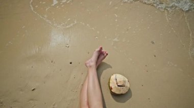 Genç bir kadın kumlu sahilde çıplak bacakları ve yumuşak deniz dalgalarının yanında taze hindistan cevizi ile rahatlıyor. Kopya alanı olan tropik tatil havası. Yüksek kalite 4k görüntü