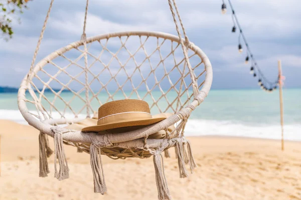Hasır şapka, kumlu tropikal plajın üzerinde asılı, turkuaz okyanus ve sicim ışıkları, huzurlu deniz kenarı rahatlama sahnesi. Yüksek kalite fotoğraf