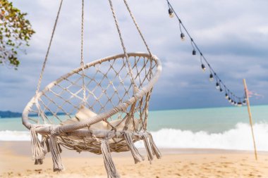 Okyanus dalgaları ve sicim ışıkları olan kumlu tropikal plajın üzerinde asılı halat sandalyesi, yaz seyahati konsepti için fotokopi alanı olan deniz kenarı rahatlama sahnesi. Yüksek kalite fotoğraf
