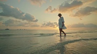 Gün batımında ve Lipa Noi plajında gün doğumunda yalın ayak yürüyen genç kadın, Koh Samui, altın gökyüzü ve geniş kopya uzayı ile huzurlu tropikal seyahat anı. Yüksek kalite 4k görüntü