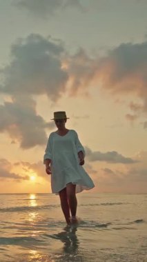 Gün batımında çıplak ayakla sığ denizde yürüyen genç bir kadın ve Lipa Noi plajında gün doğumu, Koh Samui, yumuşak ışık ve dramatik bulutlarla tropik seyahat sahnesi. Yüksek kalite 4k görüntü