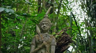 Samui 'deki Sihirli Bahçe' deki taş savaşçı heykeli yoğun tropikal ormanlarla çevrili, yosun ve eğreltiotu ile merasim bıçağı taşıyan gizemli bir ruhsal atmosfer yaratıyor. Yüksek kalite 4k görüntü