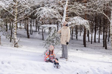 Kışın ormandaki çam ağaçlarının altında karla oynayan iki genç kız, biri sallanan bir dal, diğeri ise karlı zeminde, neşeli açık hava eğlencesi sahnesinde oturuyor. Yüksek kalite fotoğraf