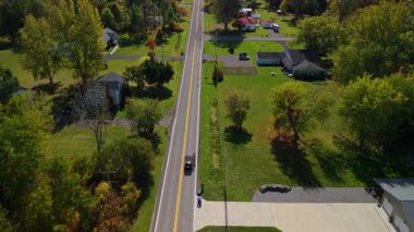 Küçük Amerikan kasabası boyunca düz bir kırsal yol boyunca giden bir arabanın hava görüntüsü, sonbahar ağaçları ve evler, insansız hava aracı perspektifi kırsal altyapı ve seyahat rotasını gösteriyor. Yüksek