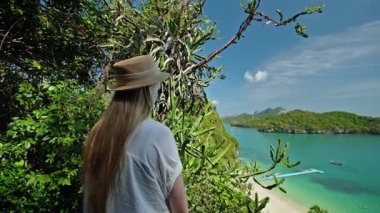Tropik bir bakış açısında duran şapkalı genç kadın, Tayland 'daki Ang Thong Deniz Parkı' ndaki zümrüt lagünü ve kireç taşı adalarına bakıyor, yemyeşil bitki ve kaktüs bitkileriyle çevrili. Yüksek