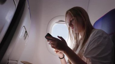 Pencerenin yanındaki uçuşta akıllı telefon kullanan genç bir kadın..