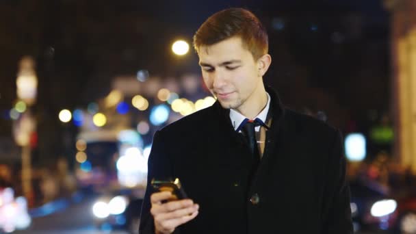 Un joven mirando la pantalla del teléfono, de pie en el fondo de la ciudad por la noche — Vídeos de Stock