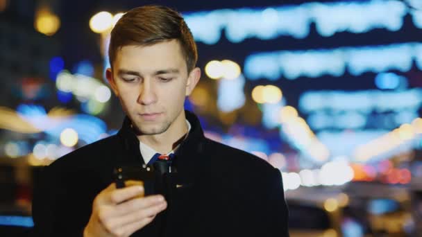 Um jovem olhando para a tela do telefone, Ele está no fundo das luzes da cidade noite — Vídeo de Stock