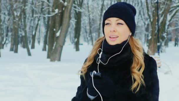 Atractiva mujer caminando en un parque de invierno, escuchando música con auriculares — Vídeo de stock