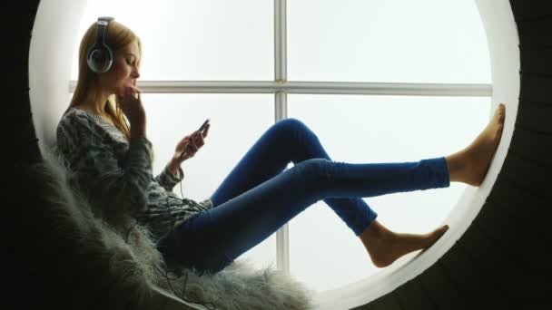 Young woman sitting at a round window and listening to music on headphones — Stock Video