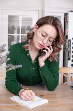 Güzel sarışın kadın, omuz hizasında saçları parlak bir odada elinde bir telefon, bir kalem ve bir defterle bir masada.