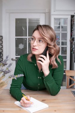 Güzel sarışın kadın, omuz hizasında saçları parlak bir odada elinde bir telefon, bir kalem ve bir defterle bir masada.