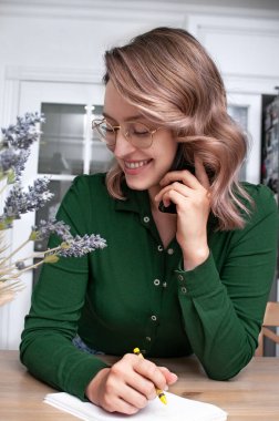 Güzel sarışın kadın, omuz hizasında saçları parlak bir odada elinde bir telefon, bir kalem ve bir defterle bir masada.