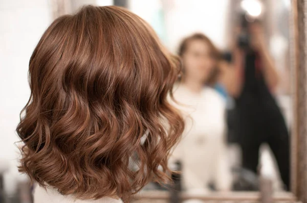 中茶色の髪の若い女性に美しい波状の髪のスタイリング室内 鏡で反射して背中からの眺め クローズアップ — ストック写真