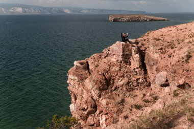 Kadın bir kayanın tepesinde oturur ve bir yaz günü Baykal Gölü manzarasına bakar.