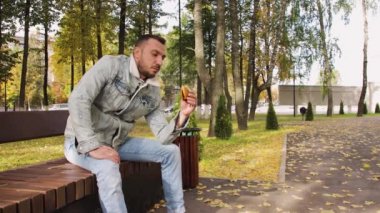 Kot elbiseli bir adam bankta oturuyor ve parkta hamburger yiyor..
