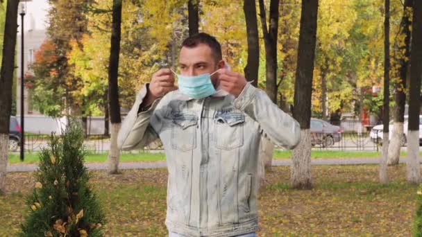 Un hombre parado en un parque se pone una máscara médica azul en la cara.. — Vídeo de stock