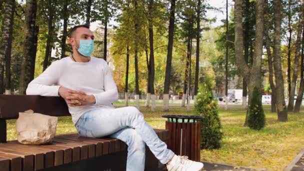 Ένας άντρας με μια ιατρική μάσκα στο πρόσωπό του κάθεται σε ένα παγκάκι στο πάρκο.. — Αρχείο Βίντεο
