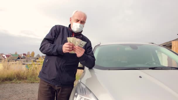 Un hombre con una máscara, apoyando sus codos en un coche, cuenta el dinero cuidadosamente. — Vídeo de stock