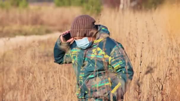 Een jongen met een medisch masker, in het hoge gras, spreekt aan de telefoon. — Stockvideo